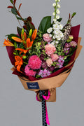 A vibrant mixed flower bouquet wrapped in brown kraft paper and red tissue, featuring pink roses, red lilies, white delphiniums, and green foliage, held by a person wearing a green sweater.