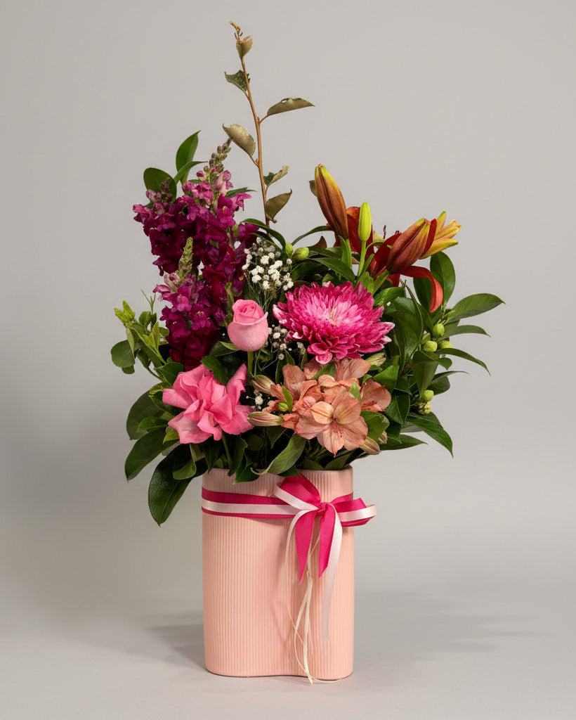 A vibrant floral arrangement in a ribbed light pink cylindrical vase, featuring pink, purple, and orange flowers with green foliage, tied with a pink ribbon, set on a wooden table in front of a dark cabinet.