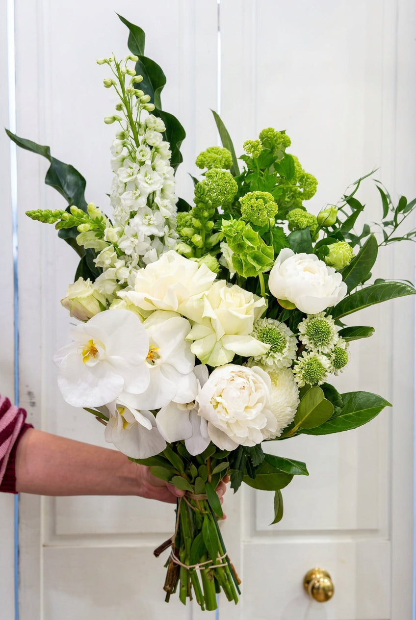 Side view of an elegant white Phalaenopsis orchid bouquet with ivory roses, green blooms and fresh foliage.